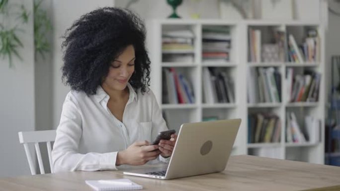 一位年轻的非洲裔美国妇女在智能手机上发短信的肖像。穿着白色上衣的卷发黑发女人坐在轻便的家庭办公室的桌