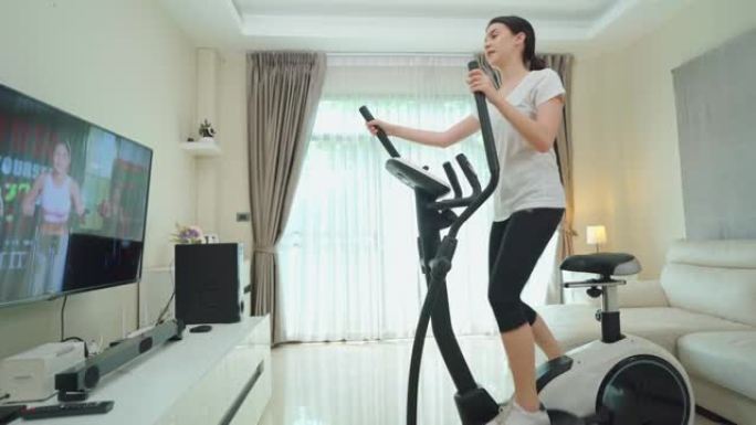 亚洲运动女性通过在家电视上观看健身现场直播或视频教程电视来锻炼。使用健身车的休闲年轻女孩在检疫过程中