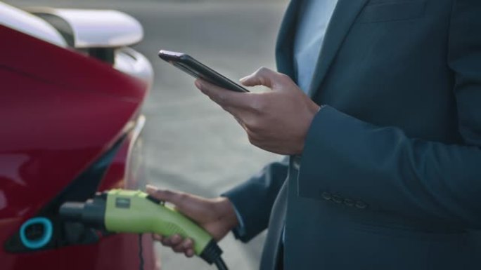 非洲男子在为现代电动汽车充电时，在智能手机上解决紧急问题。商人站在户外，靠近他的豪华红色汽车。使用智