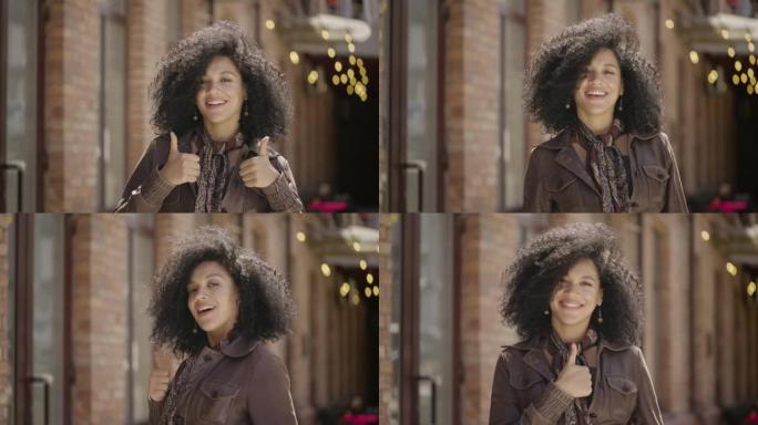 时尚的年轻非洲裔美国妇女的肖像露出竖起大拇指，手势像。穿着棕色皮夹克的黑发女人在街上对着模糊的砖砌建