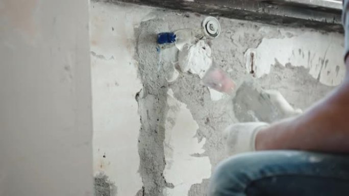 熟练的修理工在工作服上用金属抹刀，刮刀和白色抹灰膏在窗户下抹平破旧的水泥墙，用腻子粉刷平整墙面。墙壁