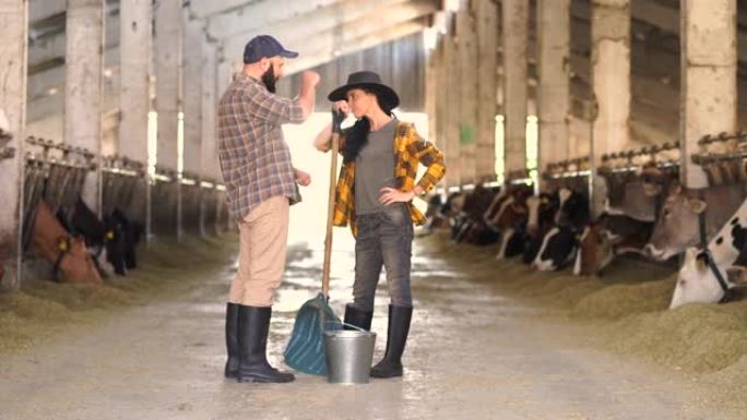 4k视频中年女农场工人戴着牛仔帽，拿着铲子，为牛棚中的年轻男工人提供建议