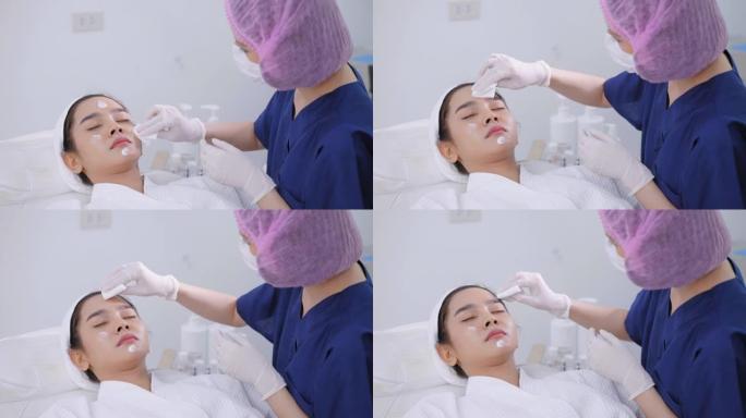 戴着卫生发帽的医务人员为年轻的亚洲妇女提供面部护理服务，清洁皮肤上的保湿霜，维生素c，穿着浴袍躺在白