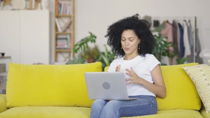 一位年轻的非洲裔美国妇女在便携式笔记本电脑上进行视频通话时的肖像。黑发，卷发，坐在明亮的家庭房间里的