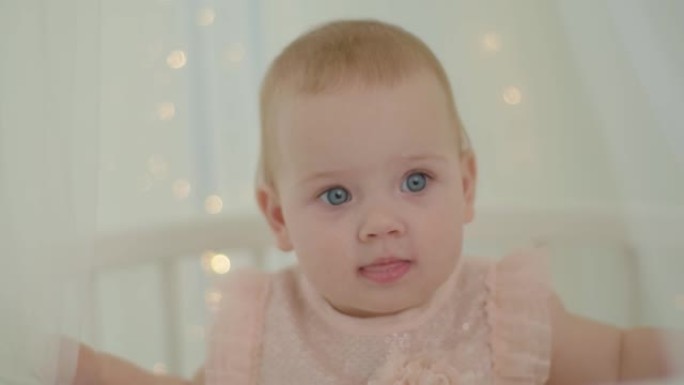 特写10个月大的蓝眼睛女婴坐在床上，带灯的顶篷。柔软明亮的婴儿室，婴儿躺在床上。