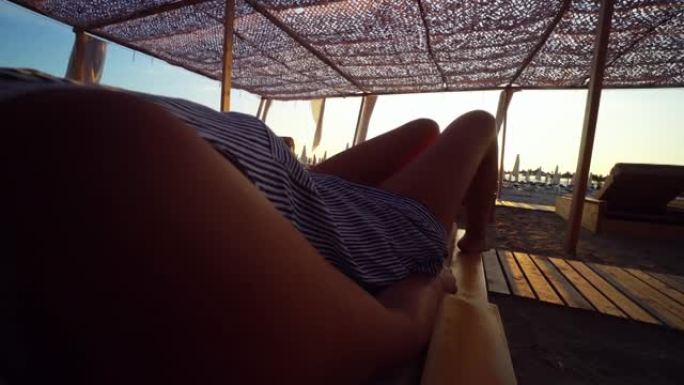 日落时，穿着衣服和帽子的晒黑女人躺在海滩上的露蒂床上躺椅上，遮阳网下