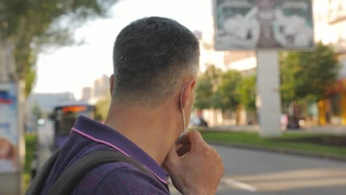 一名男子站在路边的公共汽车站等车时戴上医用口罩。