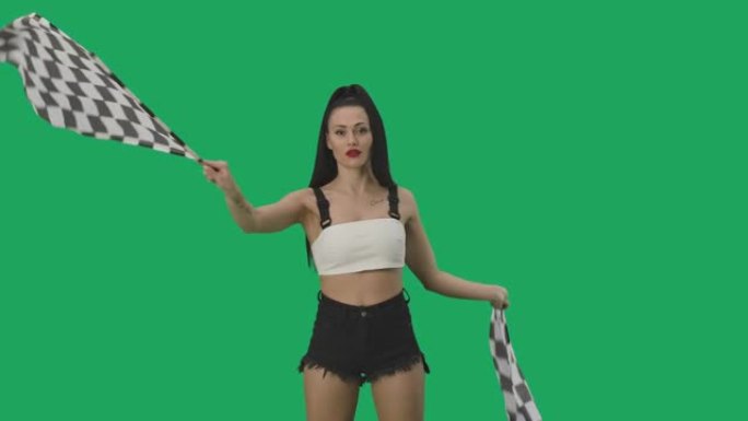 穿着短裤的性感黑发女郎挥舞着黑白方格赛车旗帜。年轻女子在摄影棚的绿屏背景下摆姿势。慢动作就绪，4k，