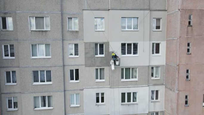 一个工业登山者悬挂在绳索上，用滚筒在建筑物的墙壁上粉刷。一名男性工人在多层建筑物的外墙上进行隔热工作