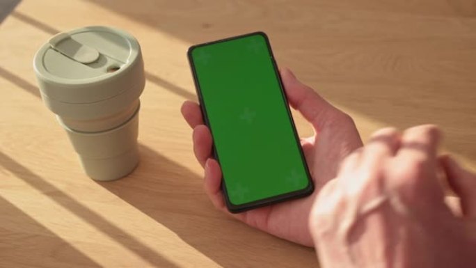 男性手拿着智能手机的垂直绿色屏幕模板，从生态可重复使用的硅胶杯中喝咖啡。手机chromakey在木桌