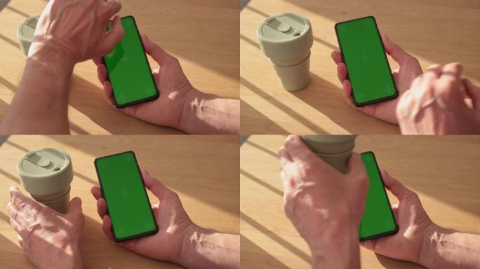 男性手拿着智能手机的垂直绿色屏幕模板，从生态可重复使用的硅胶杯中喝咖啡。手机chromakey在木桌