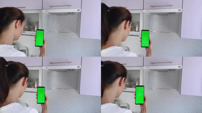 厨房里一头黑发的女人拿着一台垂直绿屏的智能手机，并阅读消息。用于广告的色度键布局。