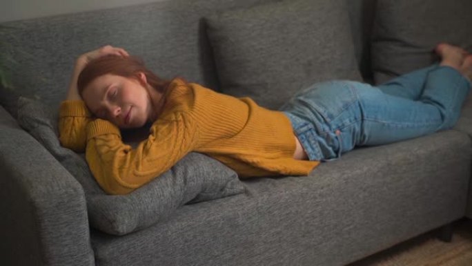 平静微笑的红发女人和安详的脸在家放松躺在舒适的沙发上享受福利的安心。