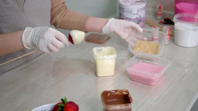 戴白手套的女孩在白巧克力和磨碎的核桃中准备草莓。