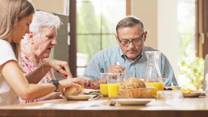 一家人正在吃早饭，父母坐在客厅里的桌子旁，他们的女儿坐在母亲旁边，他们正在准备食物，切面包，说话