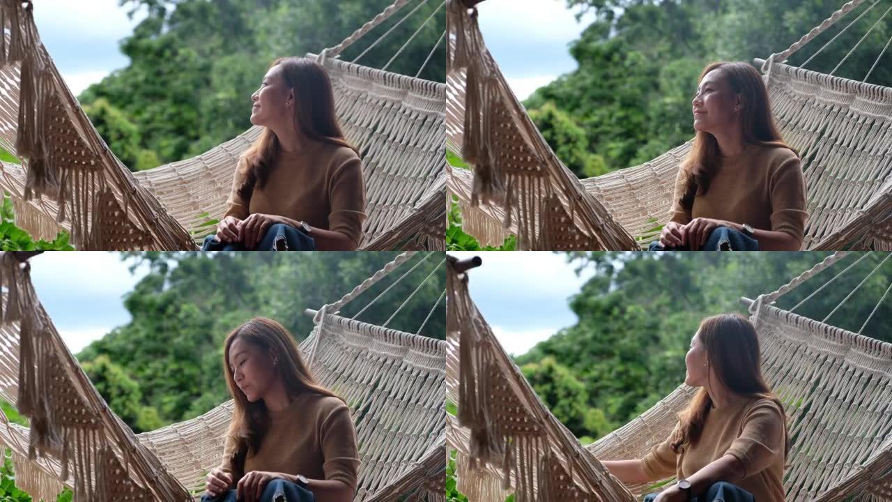 一位年轻的亚洲妇女坐在家里的吊床上放松，并享有美丽的自然风光