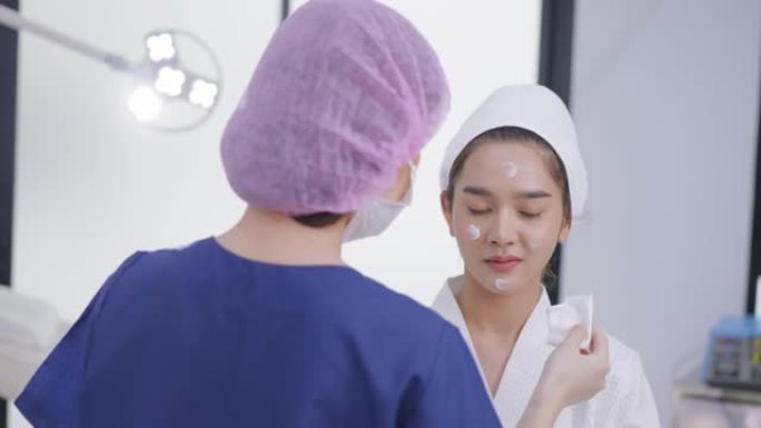 幸福的微笑医疗客户，带卫生发帽的女医生为年轻的亚洲妇女提供面部护理服务，在白色医疗沙发上穿着浴袍，在