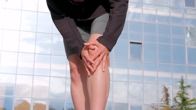 白人妇女因膝盖受伤而握住并按摩她的腿。女孩慢跑，在城市街道上锻炼; 背景建筑。疼痛的身体状况或膝盖疼