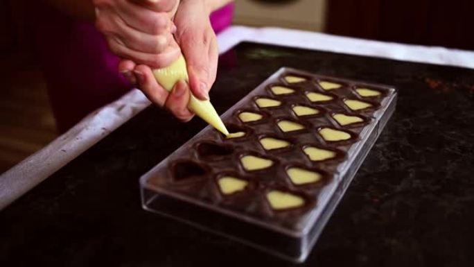 厨师糕点巧克力的特写镜头将液态奶油馅料倒入巧克力模具中，在家中准备美味的手工果子理。世界巧克力日。烹