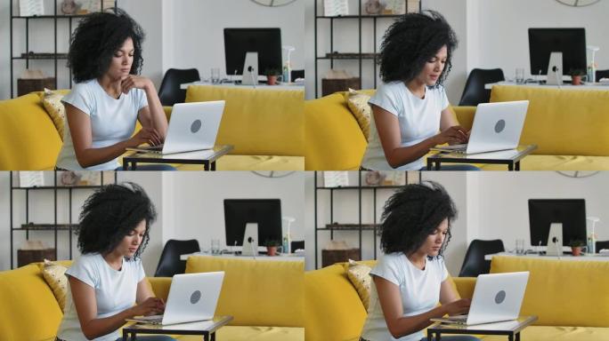 一个年轻的非洲裔美国妇女在笔记本电脑键盘上打字的肖像。黑发，卷发，坐在明亮的家庭房间里的黄色沙发上。