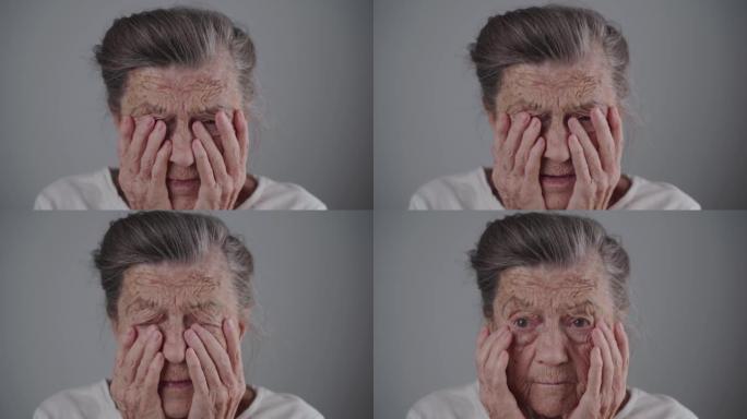 90岁高龄女性，有着深深的皱纹和白发，心理压力很大，哭着看着相机，双手放在灰色背景上。老年人的抑郁和