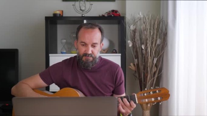 在家中的中年男子在笔记本电脑的帮助下弹吉他的前视图坐在大窗户旁边的桌子上