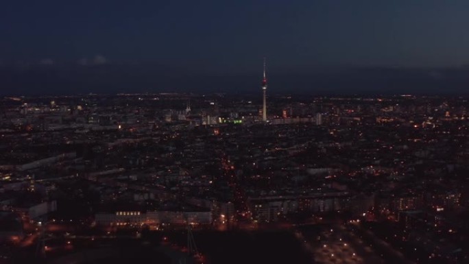 晚上飞越大城市。城市社区和城镇占主导地位的高而薄的电视塔Fernsehturm的鸟瞰图。德国柏林