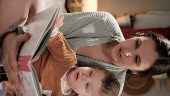 婴儿和书籍，母亲和孩子阅读。女人妈妈和2-3岁男孩的正面垂直视图晚上或晚上在卧室里穿着睡衣坐在床上，