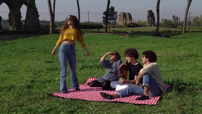 公园里无忧无虑的女人与坐在草坪上的朋友跳舞和唱歌