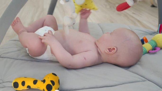 可爱的男婴抓住并拿着悬挂的玩具摇铃躺在游戏垫上，发展伸展手臂和抓住东西的能力，3个月大的新生婴儿。