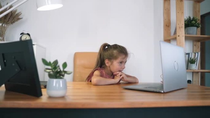 女孩看着她的笔记本电脑。孩子坐在桌旁在线交流。孩子在网上学习。家庭教育。台灯的光。在线训练自我隔离慢