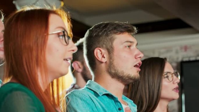 在开始体育比赛时观看国歌时，白人男女朋友站在酒吧里不动。男人和女人在酒吧唱赞美诗，手牵手。足球迷在电