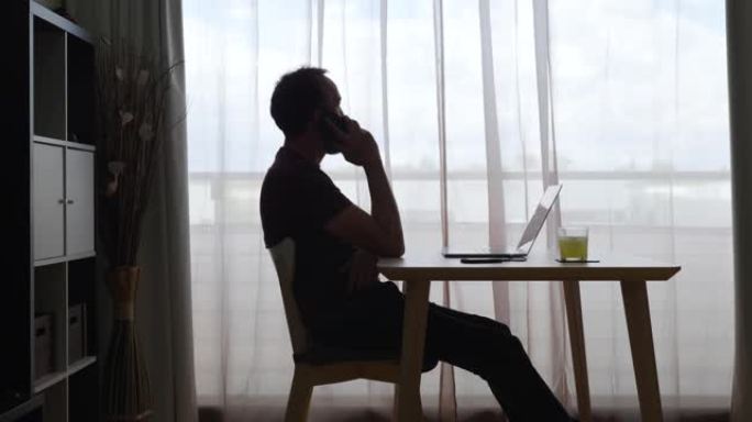 中年男子在他的背光房屋中，在笔记本电脑上坐在大窗户旁边的桌子旁，正在打电话。
