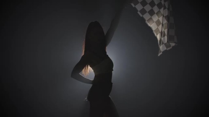 剪影的侧视图年轻女子挥舞着方格竞赛旗，标志着赛车活动的开始。黑发女人在一个带背光的黑烟工作室摆姿势。