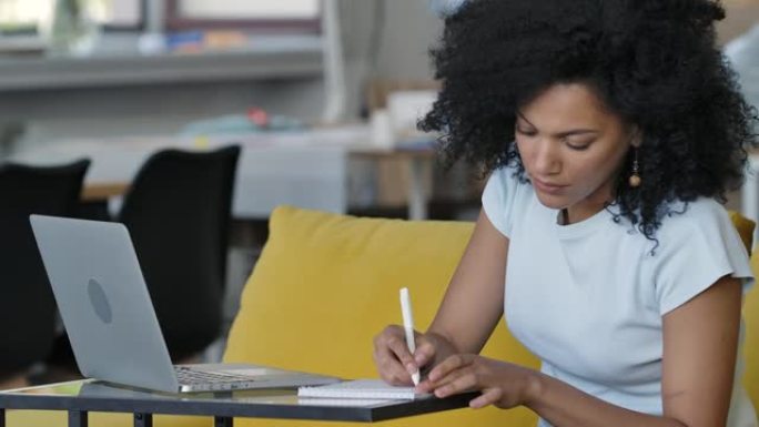 年轻的非洲裔美国妇女的肖像使用笔记本电脑在视频会议上讲话，并在笔记本电脑上做笔记。黑发卷发坐在家里的