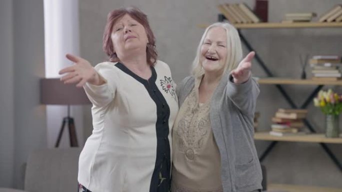 两名快乐的高级女性在镜头前亲吻空中微笑的中景。积极的女性白人朋友在客厅摆姿势。快乐退休人员在室内开会