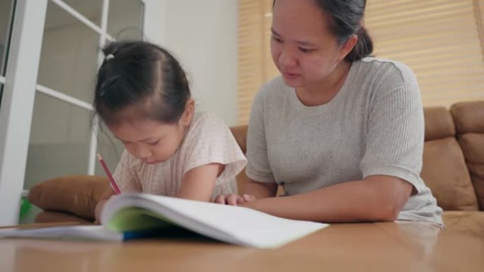 亚洲母亲照顾她的小女孩在家学习学校练习册上的写作