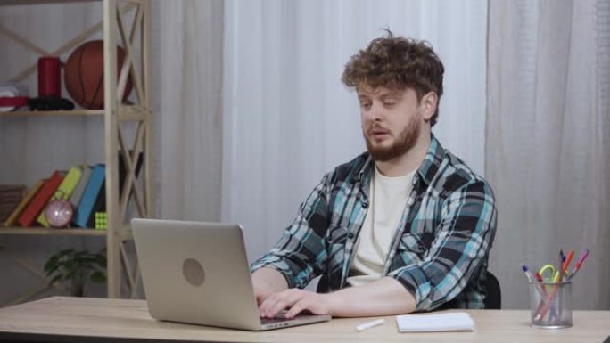 穿着方格衬衫的年轻人在笔记本电脑键盘上打字。留着胡子的红发男性坐在家庭办公室的办公桌前。特写。慢动作