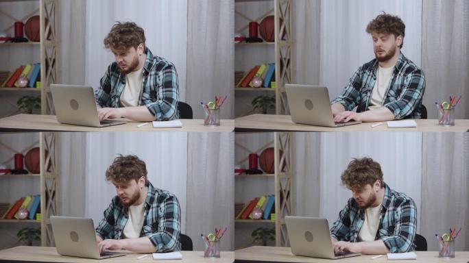 穿着方格衬衫的年轻人在笔记本电脑键盘上打字。留着胡子的红发男性坐在家庭办公室的办公桌前。特写。慢动作