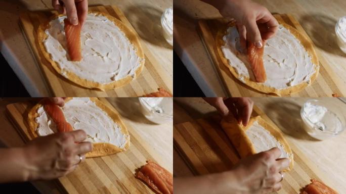 薄薄的美味彩色煎饼。女人的手将鲑鱼片放在木板上的黄色煎饼上，上面涂有奶油奶酪。滚动煎饼。4k视频。