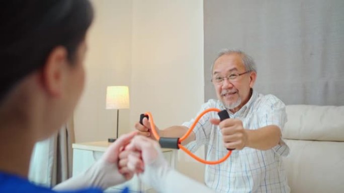亚洲残疾老年男子坐在沙发上，在治疗师护士的支持下做理疗师。老年残疾人在家庭护理中使用阻力拉伸带运动对