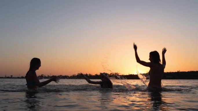 穿着比基尼的快乐湿女孩剪影在日落时互相泼水很有趣。女性朋友在日出时在池塘里飞溅很有趣。女人在黄昏去湖
