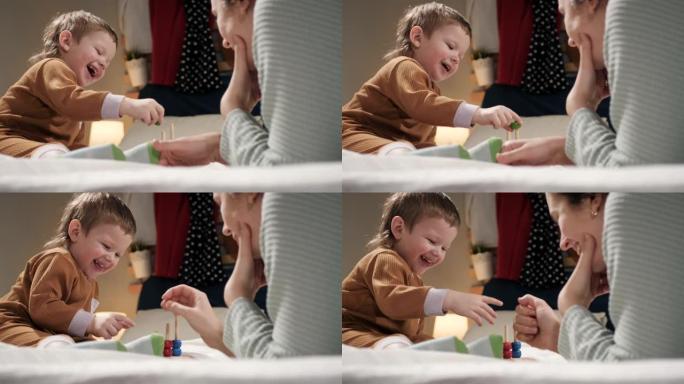 妈妈和宝宝在玩。从母亲的肩膀后面看2-3岁的小快乐笑男孩，他们晚上躺在床上的睡衣上，玩益智玩具。慢动