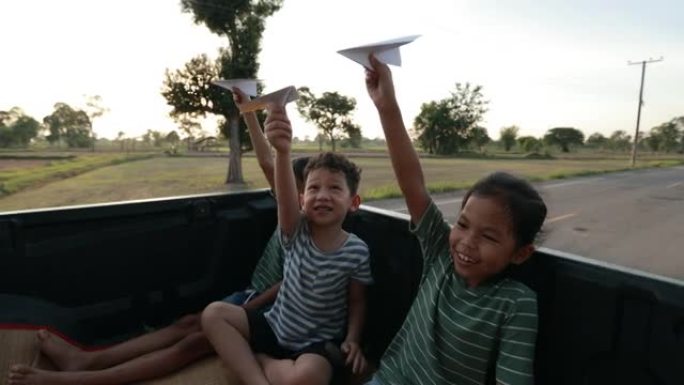 三个亚洲兄弟姐妹男孩和女孩坐在汽车后备箱里，在乡下玩玩具纸飞机。