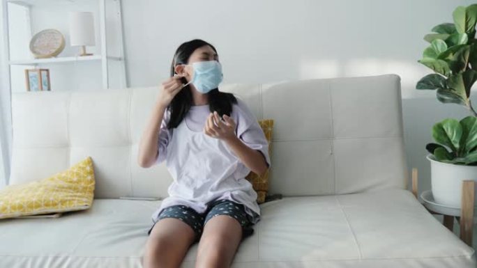亚洲女孩坐在沙发附近的窗户上戴着口罩在家，新型冠状病毒肺炎期间的医疗保健生活方式概念。
