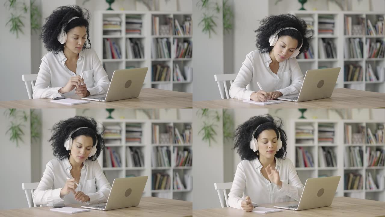 使用笔记本电脑和耳机在记事本上做笔记的非裔美国妇女在视频电话会议上讲话的肖像。布鲁内特坐在家庭办公室