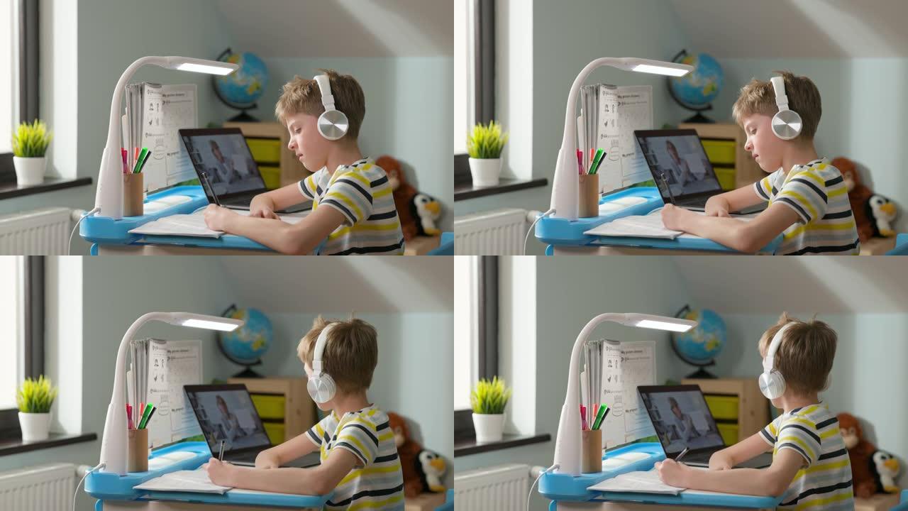 男孩坐在家里的笔记本电脑上。在线课程。戴着耳机的孩子听在线课程，然后在笔记本上写字。