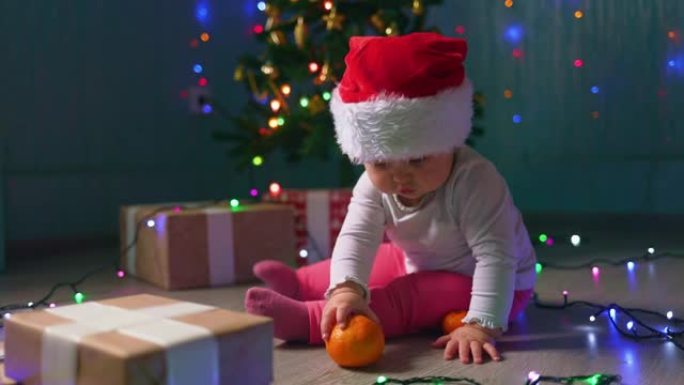 戴着圣诞帽的可爱的蹒跚学步的女孩坐在圣诞树附近，吃橘子。慢动作。冬季季节性假期的概念