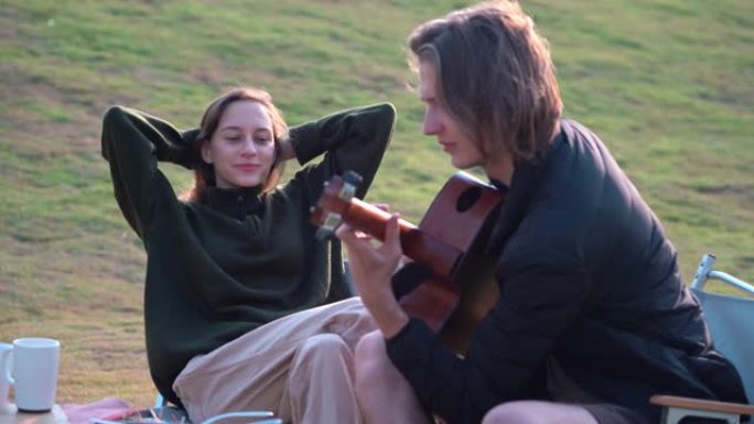 年轻的时髦夫妇弹吉他，早上在大自然公园的草原乡村一起在干草上唱一首歌，户外有阳光露营