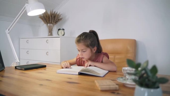 一个女孩坐在桌子旁看书。这孩子正在学习阅读。家庭教育。一个聪明的孩子。看书。儿童童话。自我隔离。慢动